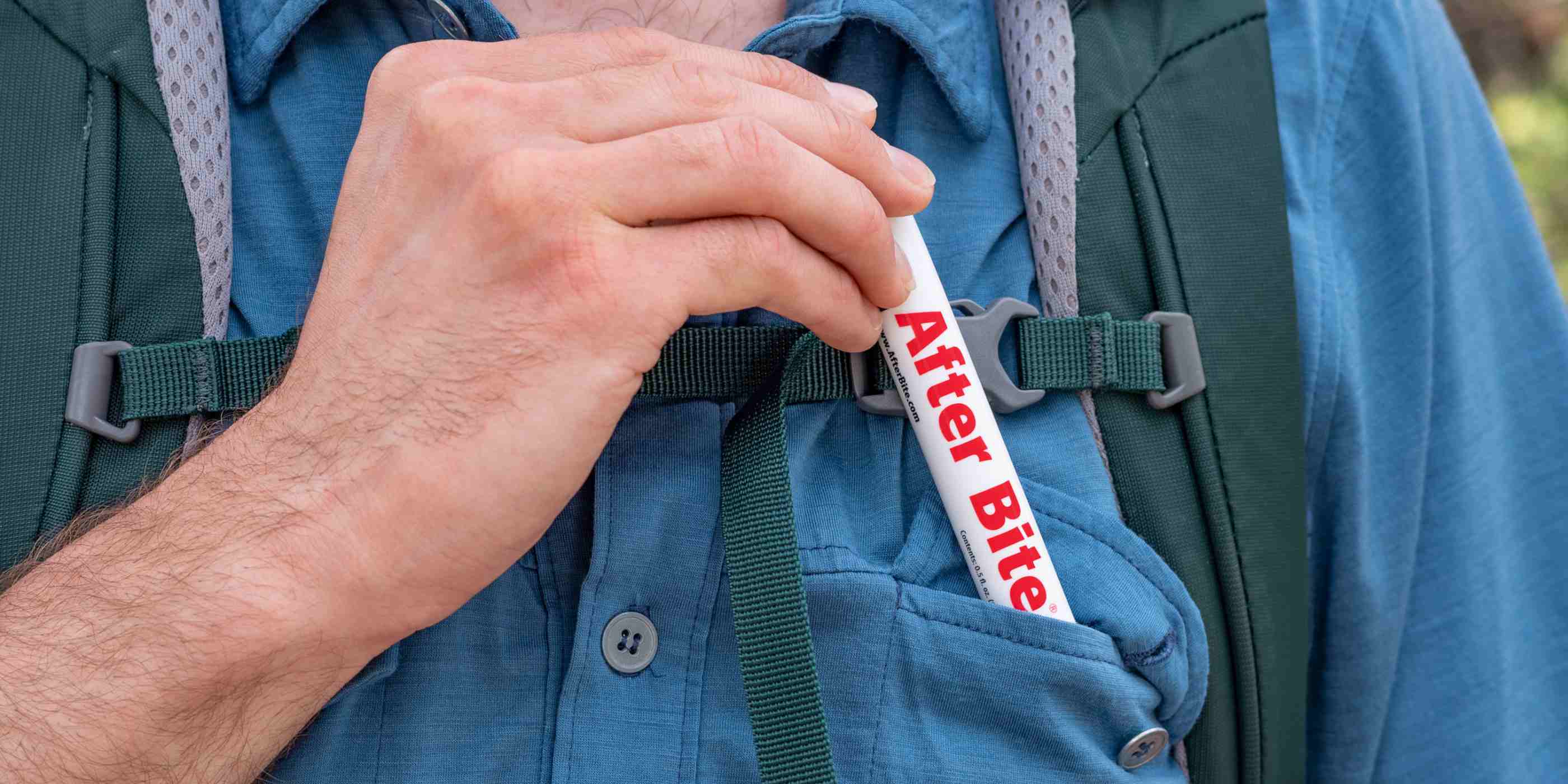Person in blue button up shirt with green backpack pulling tube of After Bite out of shirt pocket