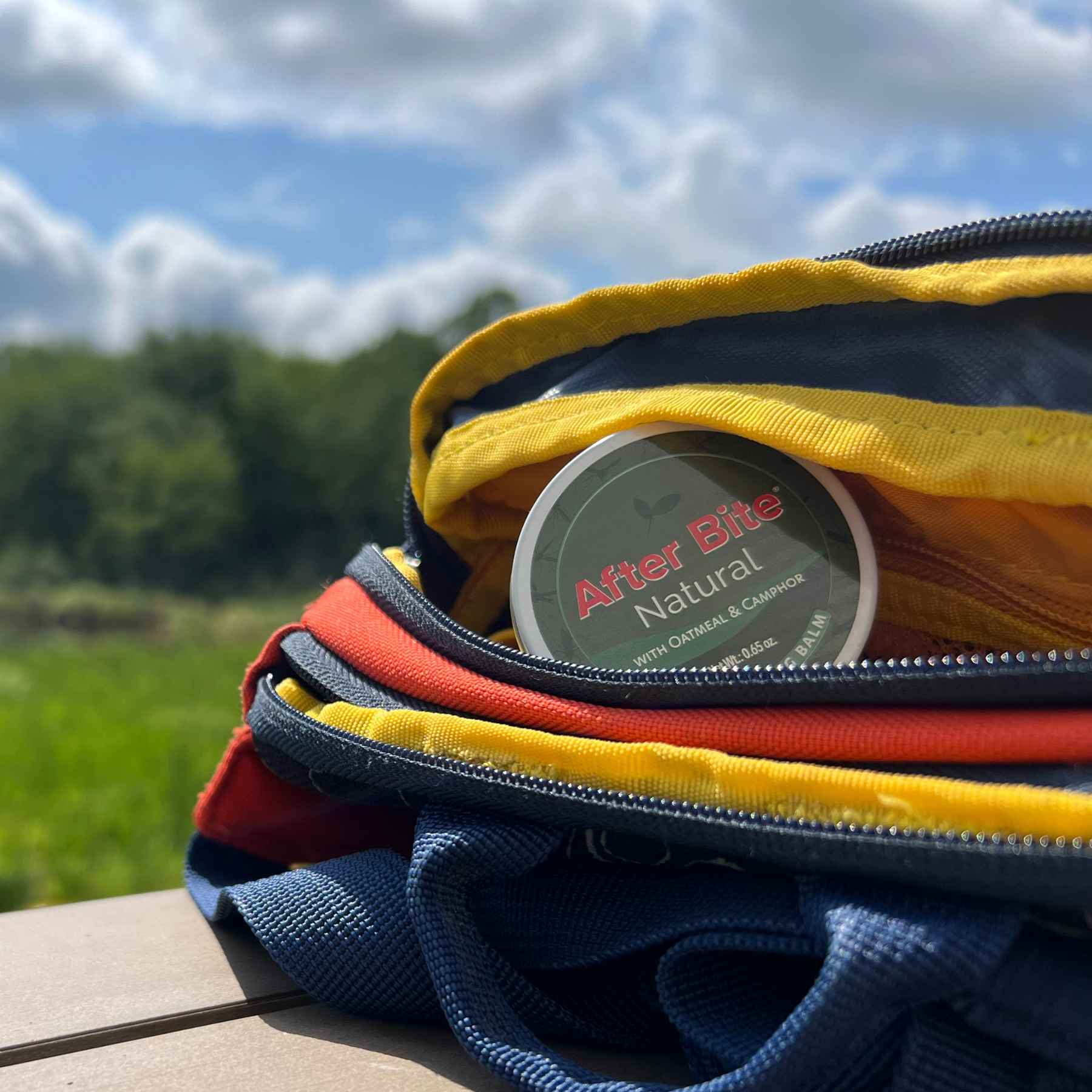 Tin of After Bite Natural peeking out of blue, red, and yellow backpack on a picnic table in summer