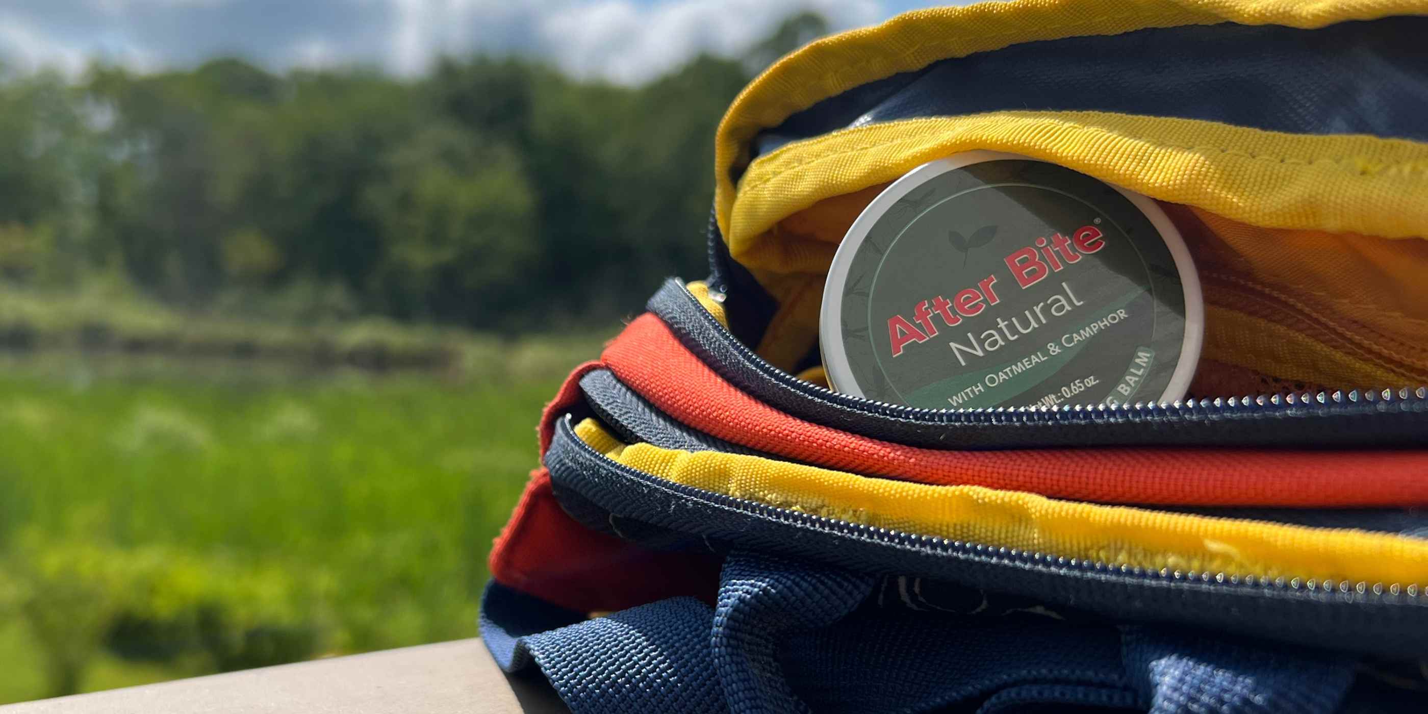 Tin of After Bite Natural peeking out of blue, red, and yellow backpack on a picnic table in summer