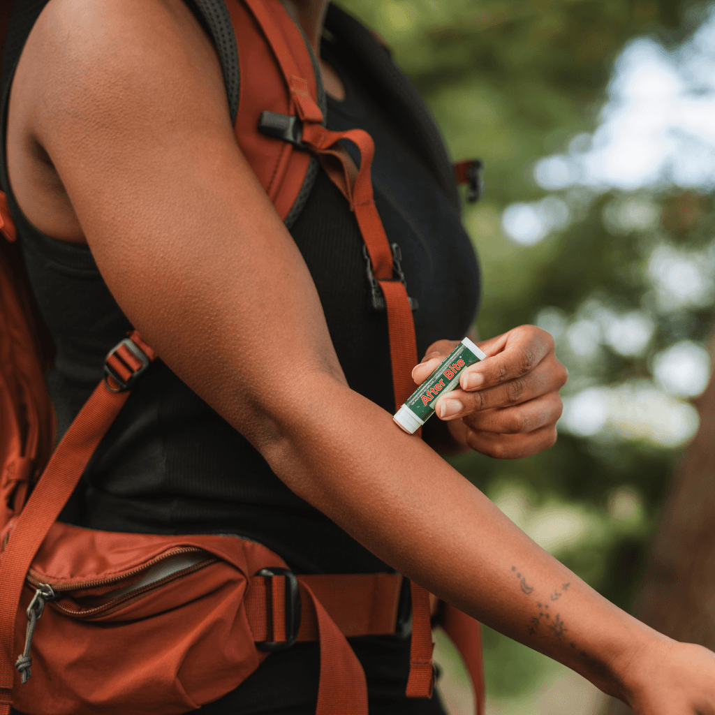 Woman applying the After Bite Natural stick to her arm while wearing an orange backpack